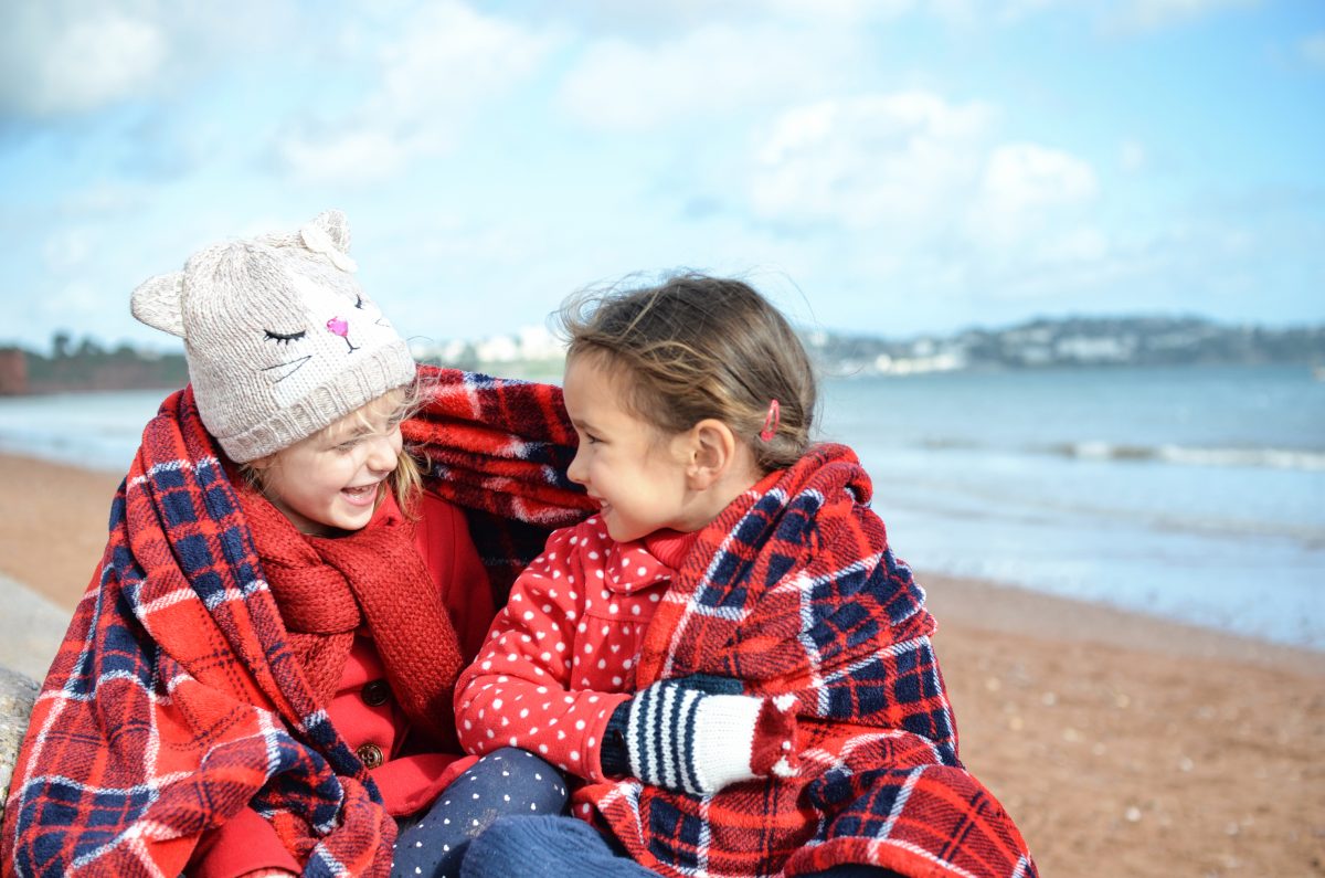 Children wrapped up in a blanket with hats and gloves on a winters day at the beach