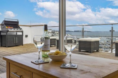Spacious holiday home balcony with stunning sea views, outdoor seating, BBQ, and wine glasses set for al fresco dining